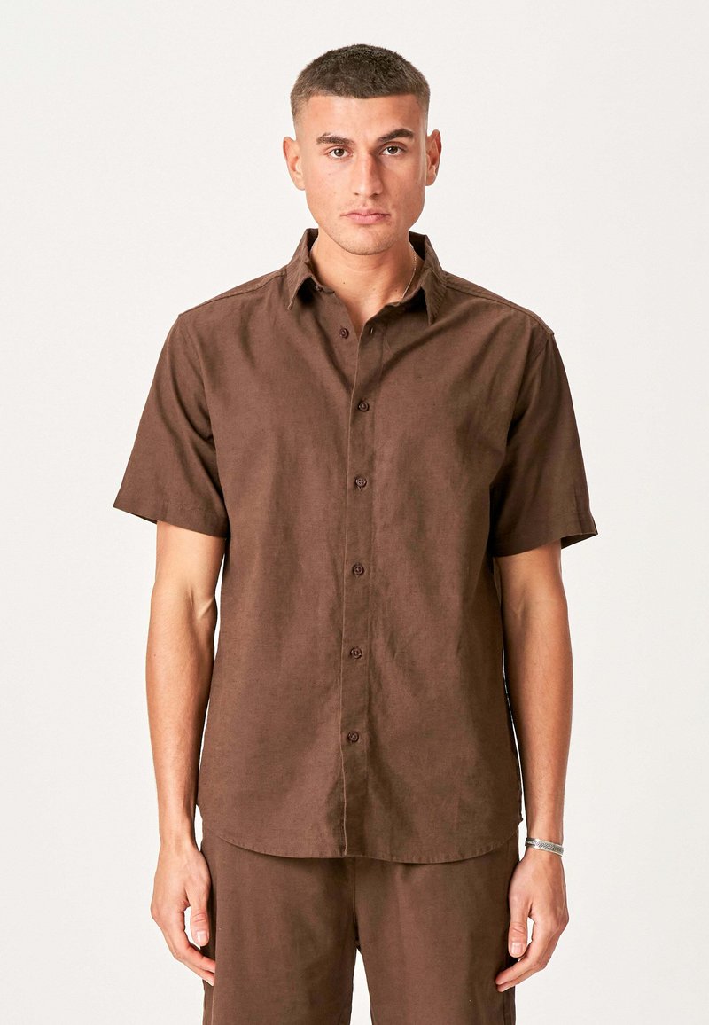 Mand står med fronten mod kameraet iført en brun kortærmet skjorte med knapper og matchende brune bukser foran en ensfarvet baggrund.