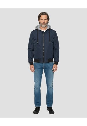 Granatowa kurtka bombowiec z szarym kapturem, zamkiem błyskawicznym z przodu i czarnymi ściągaczami na rękawach. Noszona z niebieskimi dżinsami i czarnymi butami.