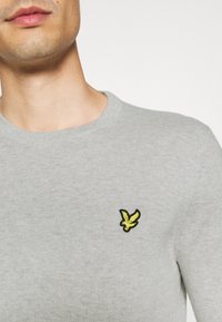 Grijze gebreide sweatshirt met een ronde halslijn en een gele geborduurde vogel logo op de linkerborst. Soepele textuur, casual ontwerp.