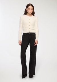 Cardigan bianco peloso con scollatura rotonda e sei bottoni in perla, abbinato a jeans neri a vita alta e gamba larga con tacchi neri.