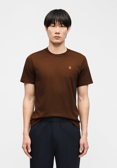 Jeune homme aux cheveux noirs et raides portant un t-shirt marron uni à manches courtes et un pantalon foncé, debout devant un fond clair.