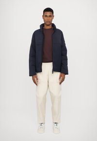 Jachetă puffer în navy cu guler înalt, fermoar frontal și două buzunare pe piept, asociată cu pantaloni cargo bej și adidași albi. Textură netedă.