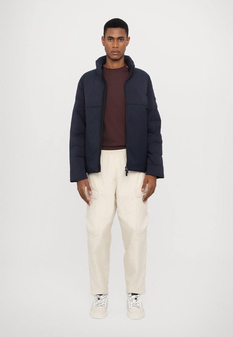 Marineblauer Pufferjacke mit hohem Kragen, Frontreißverschluss und zwei Brusttaschen, kombiniert mit beigen Cargo-Hosen und weißen Sneakern. Glatte Textur.