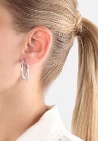 Pendiente de aro de plata con una textura suave y una forma ligeramente ovalada, colgando de la lóbulos de la oreja sobre un fondo claro.