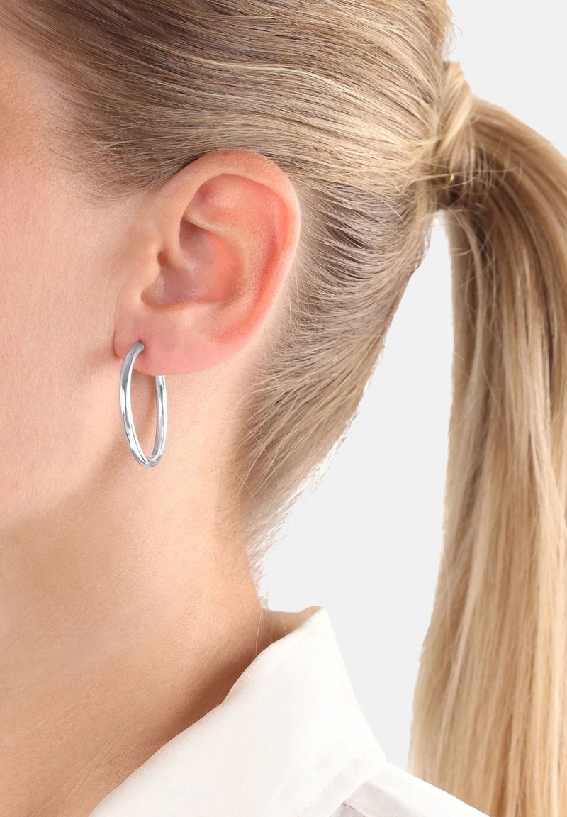 Pendiente de aro de plata con una textura suave y una forma ligeramente ovalada, colgando de la lóbulos de la oreja sobre un fondo claro.