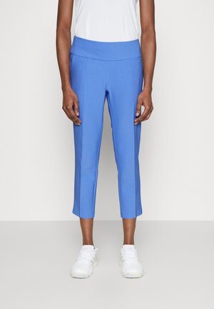 Osoba ubrana w niebieskie, krótkie, dopasowane spodnie i białe trampki, stojąca na jasnym, jednolitym tle.