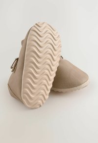 Beige ruskindsko med en struktureret gummisål med en bølget mønster, sidelukning med spænde og en rund tådesign.
