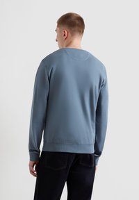 Felpa azzurra chiara con colletto rotondo e polsini a coste. Tessuto liscio, vestibilità rilassata. Abbinata a jeans scuri, vista da dietro.