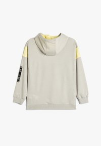 Lichtgrijze hoodie met een gele accent op de schouder, ribgebreide manchetten en zoom, platte naden en een klein logopatch op de mouw.