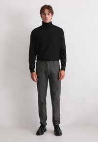 Junger Mann in einem schwarzen Rollkragenpullover, grauen Anzughosen und schwarzen eleganten Schuhen, der vor einer schlichten weißen Wand steht.