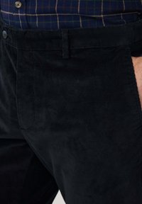 Pantalons en velours côtelé noir avec une surface texturée, dotés d'une fermeture à bouton et de poches latérales, montrés partiellement avec une chemise à carreaux en dessous.