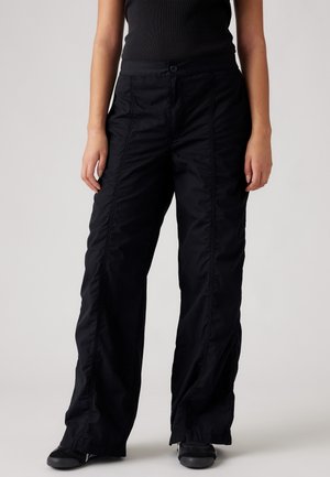 Persona con pantalones negros de corte holgado con costuras verticales visibles y zapatos negros, de pie frente a un fondo neutro.