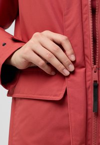 Veste rouge résistant à l'eau avec une texture lisse, dotée d'une fermeture éclair, d'un rabat de poche latérale et d'accents en matériel noir contrasté.