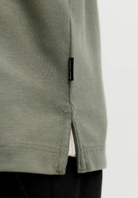 Gros plan sur l'ourlet latéral d'une chemise en tissu vert avec une étiquette noire portant la mention "Jack & Jones" et un petit détail fendu au-dessus d'un pantalon noir.