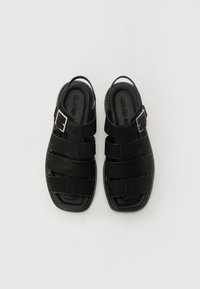 Sandalias de cuero negro con diseño de parte superior tejida, hebilla ajustable y suela plana. Acabado suave y forma rectangular con costuras en contraste.