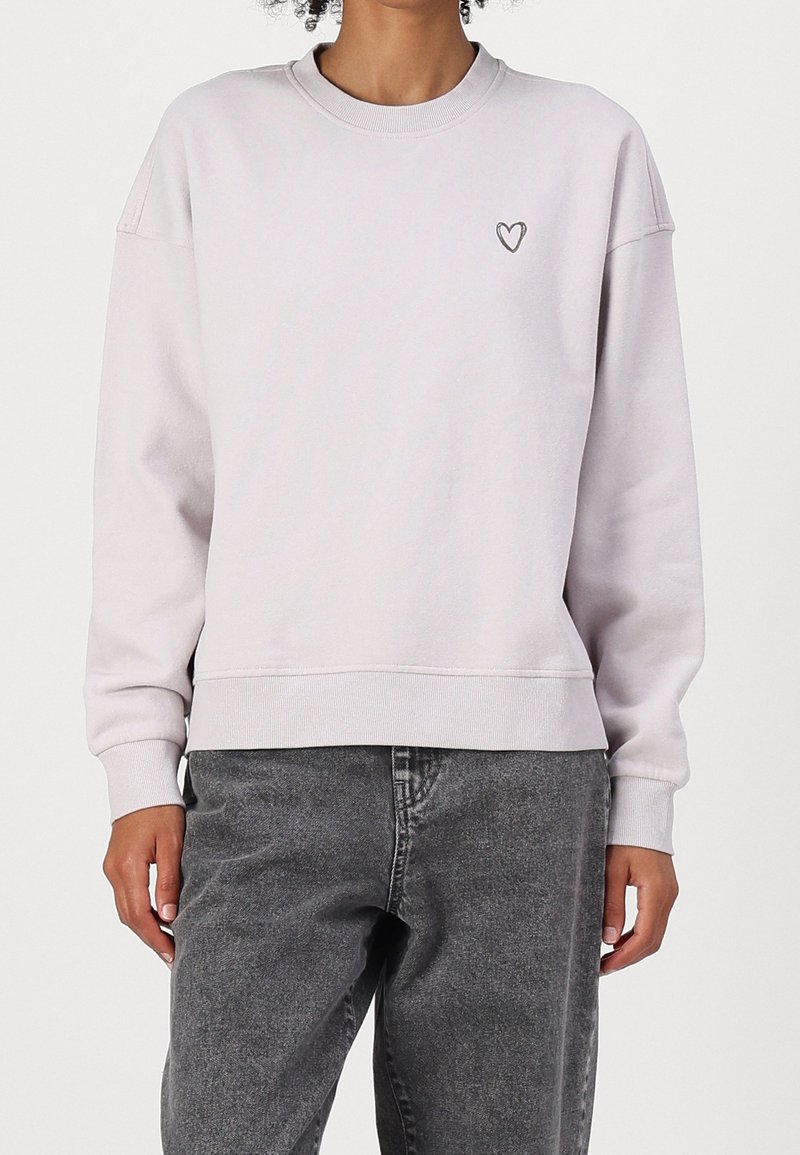 Personne portant un sweat-shirt violet clair avec un petit motif de cœur sur le côté gauche de la poitrine et un jean noir délavé, debout devant un fond uni.