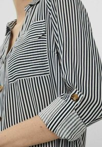 Gros plan sur une personne portant une chemise rayée verticalement en noir et blanc avec une poche et une manche retroussée fixée par un bouton marron.