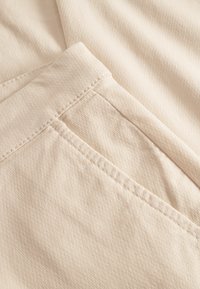 Beige textiel met een fijne structuur, waarop een close-up te zien is van naden en een pochet detail. De stof heeft een subtiel, fijn patroon en een zachte afwerking.