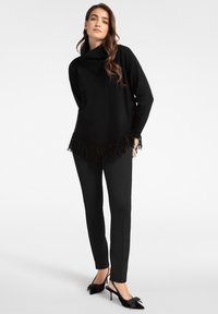 Svart stickad tröja med luftig krage och fransdetaljer vid fållen, kombinerad med svarta smala byxor och svarta klackskor med rosetter.