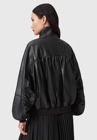 Černá kožená bombardérka s vysokým límcem, nařaseným zadním dílem a žebrovaným lemem. Disponuje měkkou, lesklou texturou a objemovými rukávy.