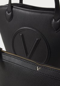 Black tote bag made of textured leather with a prominent embossed logo. Features a zipper closure and gold hardware accents.