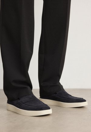 Zapatos navy de ante sin cordones con una suela de goma crema, que presentan un diseño cosido y una parte superior texturizada, combinados con pantalones negros.