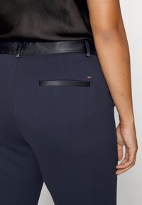 Marineblaue Hose mit glatter Textur, ausgestattet mit einem schwarzen Lederbund und einer einzelnen Gesäßtasche mit horizontalem Lederbesatz.