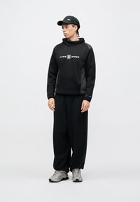 Μαύρο φούτερ με κουκούλα και λογότυπο, συνδυασμένο με φαρδύ μαύρο παντελόνι και ανοιχτό γκρι αθλητικά παπούτσια. Η καπέλο είναι επίσης μαύρο, με λογότυπο.