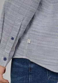 Gestreept shirt van lichtgewicht stof, met blauwe knopen, een zijkraaglabel en detail bij de manchetten. Close-up toont de textuur en het patroon.