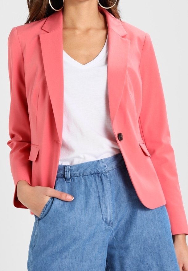 Femme portant un blazer rose corail, un t-shirt blanc à col en V, un pantalon taille haute bleu et de grandes boucles d'oreilles créoles, la main dans la poche.