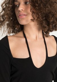 Blusa preta de manga longa com um decote profundo e cortes largos nos ombros, apresentando um tecido texturizado e detalhes em tiras finas.