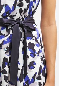 Détail d'une robe à fleurs avec une base blanche et des motifs audacieux en bleu, noir et gris ; elle comporte une ceinture en satin bleu foncé nouée en un nœud.