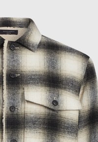Chaqueta de cuadros en blanco y negro, con forro de sherpa, botones y cuello puntiagudo, además de un bolsillo de solapa en el pecho.
