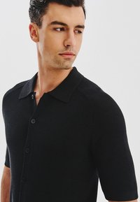 Mężczyzna w czarnej koszuli z krótkim rękawem z guzikami, patrzący w lewo na jednolity jasny tle.