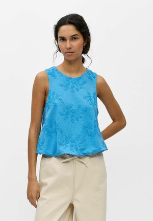 Femme aux cheveux foncés portant un haut bleu sans manches avec broderies florales et un pantalon beige clair sur fond blanc uni.