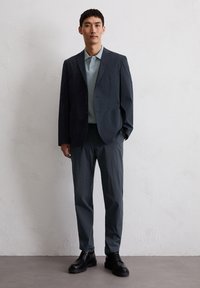 Granatowa marynarka na jasnoniebieskim podkoszulku, w zestawieniu z ciemnoszarymi spodniami i czarnymi sznurowanymi butami, na teksturowanym jasnym tle.