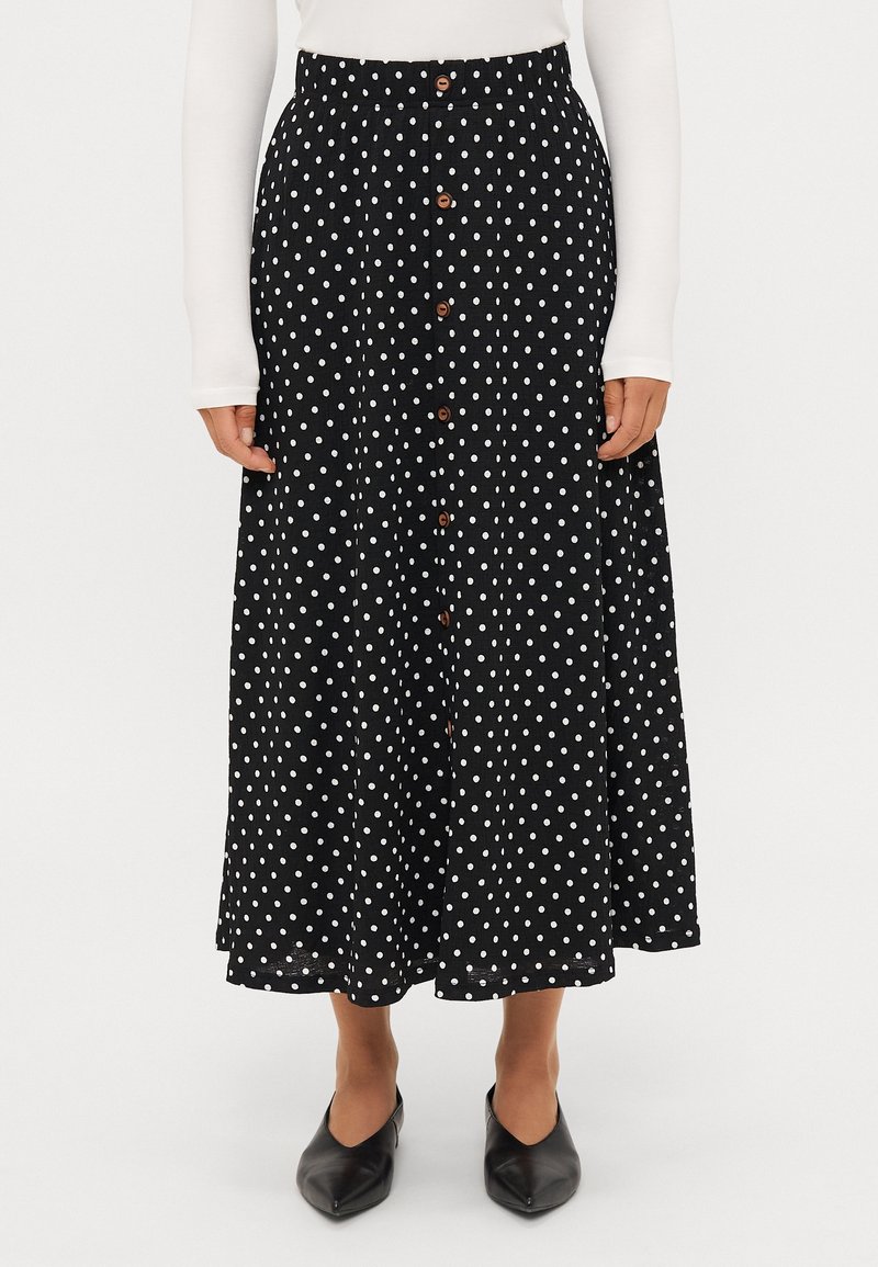 Jupe midi noire à pois blancs, avec des boutons marron sur le devant, portée avec un haut blanc à manches longues et des chaussures noires à bouts pointus.