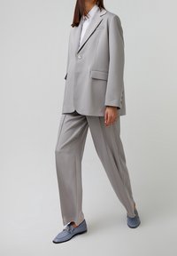 Grau maßgeschneiderter Blazer und Hose aus glattem Stoff. Der Blazer hat einen einzigen Knopf und Fronttaschen. Kombiniert mit blauen Loafers.