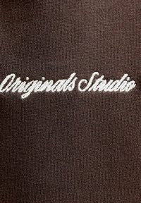 Texte blanc en cursive « Originals Studio » brodé sur un tissu tricoté marron foncé.