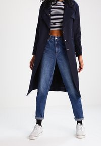 Kvinna i marinblå trenchcoat, svart och vit randig crop top, högmidjade upprullade jeans, svarta strumpor och vita sneakers.