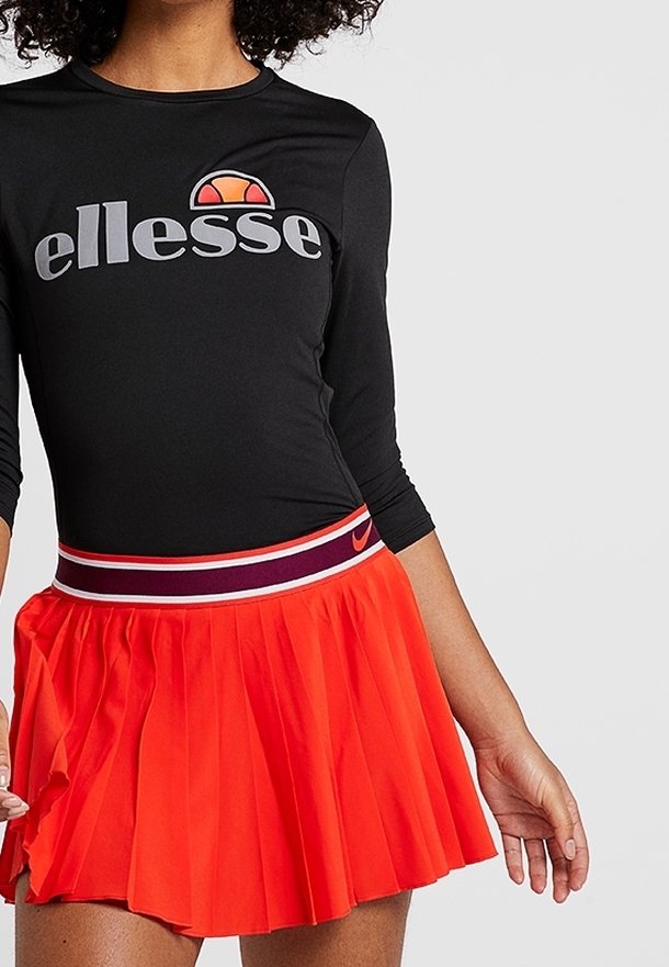 Czarny top z długim rękawem z logo "ellesse", zestawiony z plisowaną pomarańczową spódnicą z granatowym pasem i białymi akcentami.