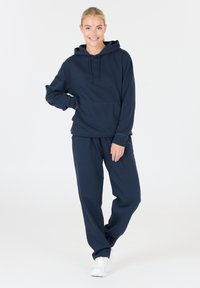 Sudadera con capucha azul marino, con un bolsillo en la parte frontal y cordones, combinada con pantalones de chándal a juego, ambos de un tejido suave y texturizado. Zapatillas blancas.