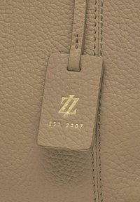 Sac en cuir texturé beige avec coutures, et étiquette en cuir rectangulaire attachée, embossée du logo doré et du texte "EST. 2007".