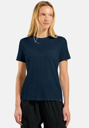 Femme aux longs cheveux blonds portant une chemise à manches courtes bleu marine et un pantalon noir, debout devant un fond clair.