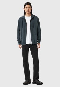 Felpa con cappuccio a zip di colore verde scuro, realizzata in tessuto morbido. Abbinata a jeans neri e scarpe nere robuste. Design minimalista e accenti sottili.