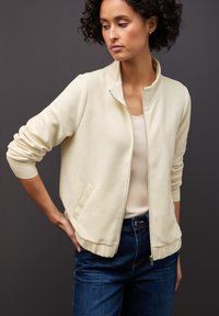 Giacca zip con texture a coste di colore crema chiaro, colletto alto e due tasche laterali, abbinata a una maglietta leggera e jeans scuri.