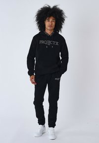 Sudadera de algodón negra con cordones y bolsillo delantero, que presenta el logo en blanco "PROJECT X PARIS". Combinada con pantalones jogger negros a juego y zapatillas blancas.