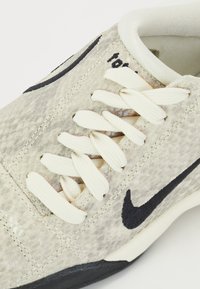 Zapatillas gris claro con parte superior texturizada, cordones blancos, swoosh negro de Nike y suela de goma negra. Detalles acentuados en los laterales.
