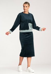 Zestaw loungewear z aksamitu, zawierający ciemnozieloną bluzkę z jasnzieloną kieszonką i mankietami, w połączeniu z pasującą długą spódnicą i białymi trampkami.