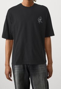 Svart bomullströja med korta ärmar, prydd med en liten vit blommig broderi på vänster bröst. Enkel rund halsringning och tyg som sitter löst.